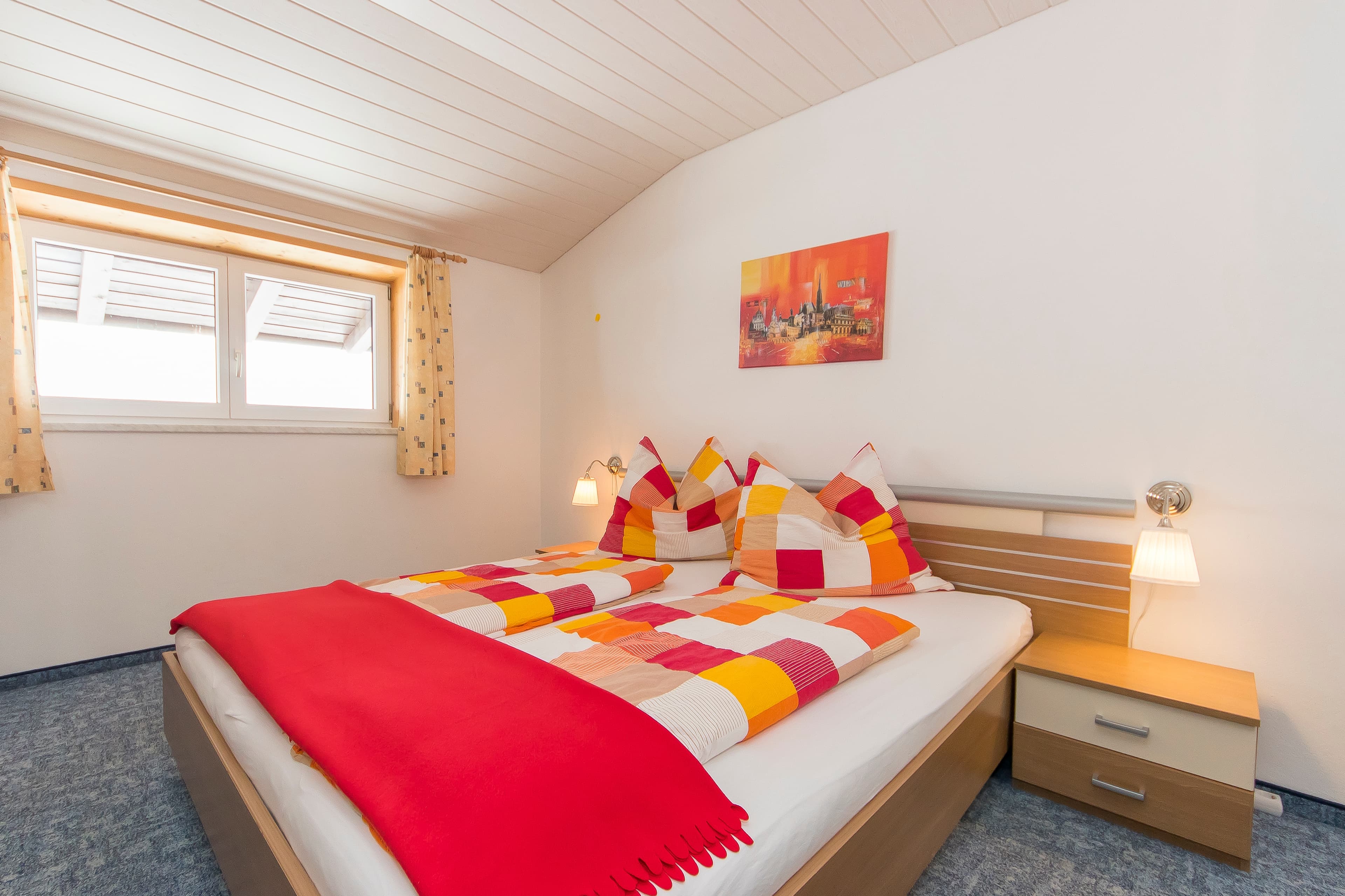 Hinterbichlhof · Apartment Rosengarten - A bedroom with a sloped wooden ceiling and white walls, featuring a double bed with red and colorful patterned bedding. A mirrored wardrobe is on the left. A window with patterned curtains allows natural light to enter. A painting hangs on the wall above the bed.